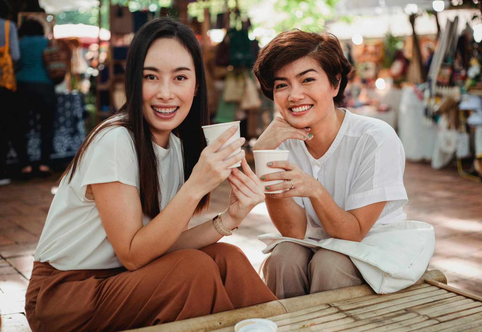 Optimistic attractive Asian females in stylish summer clothes sitting at table in outdoor cafeteria with coffee to go and looking at camera with charming smiles
